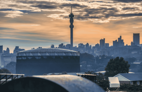 Johannesburg city skyline at sunset with the Hillbrow Tower prominent in the center, silhouetted buildings, and dramatic clouds in the sky.