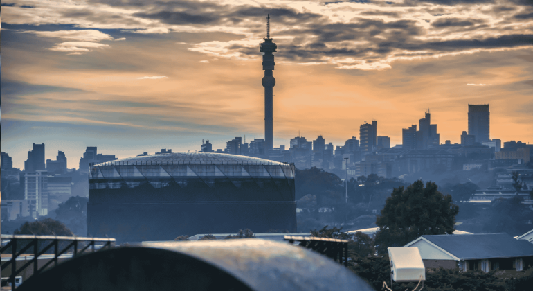 Johannesburg city skyline at sunset with the Hillbrow Tower prominent in the center, silhouetted buildings, and dramatic clouds in the sky.
