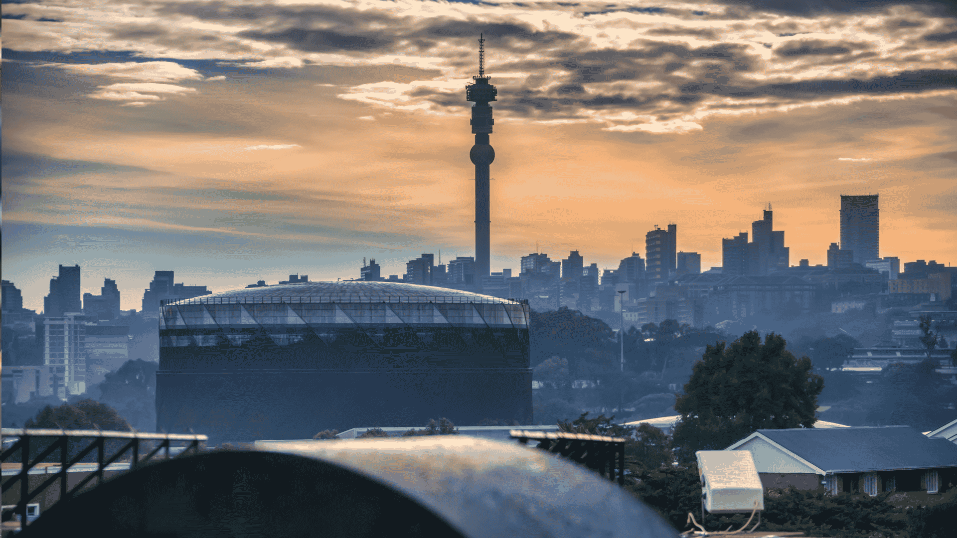 Johannesburg city skyline at sunset with the Hillbrow Tower prominent in the center, silhouetted buildings, and dramatic clouds in the sky.