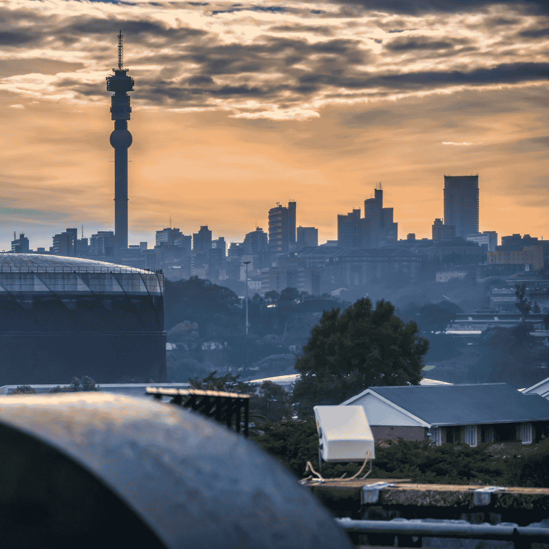 Johannesburg city skyline at sunset, featuring tall buildings, the Hillbrow Tower, and cloudy skies with an orange glow. Rooftops and trees are visible in the foreground.