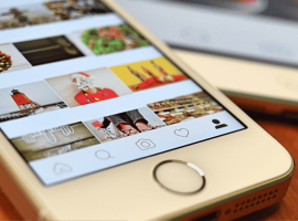 A close-up of a smartphone displaying a social media photo gallery with holiday-themed images, including wreaths, Santa, and a lighthouse. Another phone is partially visible in the background on a wooden surface.