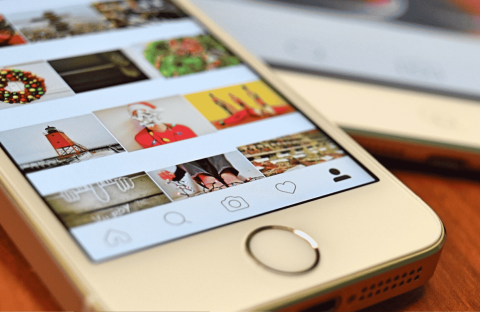 A close-up of a smartphone displaying a social media photo gallery with holiday-themed images, including wreaths, Santa, and a lighthouse. Another phone is partially visible in the background on a wooden surface.
