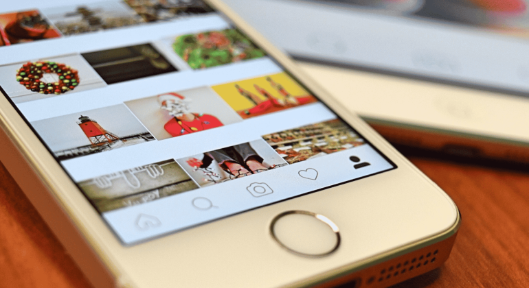 A close-up of a smartphone displaying a social media photo gallery with holiday-themed images, including wreaths, Santa, and a lighthouse. Another phone is partially visible in the background on a wooden surface.