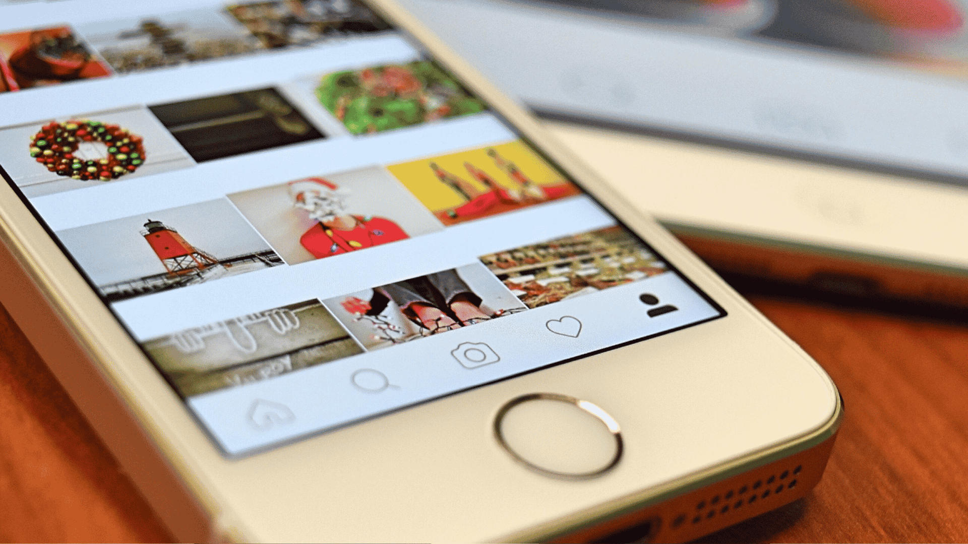 A close-up of a smartphone displaying a social media photo gallery with holiday-themed images, including wreaths, Santa, and a lighthouse. Another phone is partially visible in the background on a wooden surface.