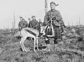 A soldier in a kilt stands beside an antelope on a barren field, with other soldiers in the background. The scene appears historical, possibly from wartime.