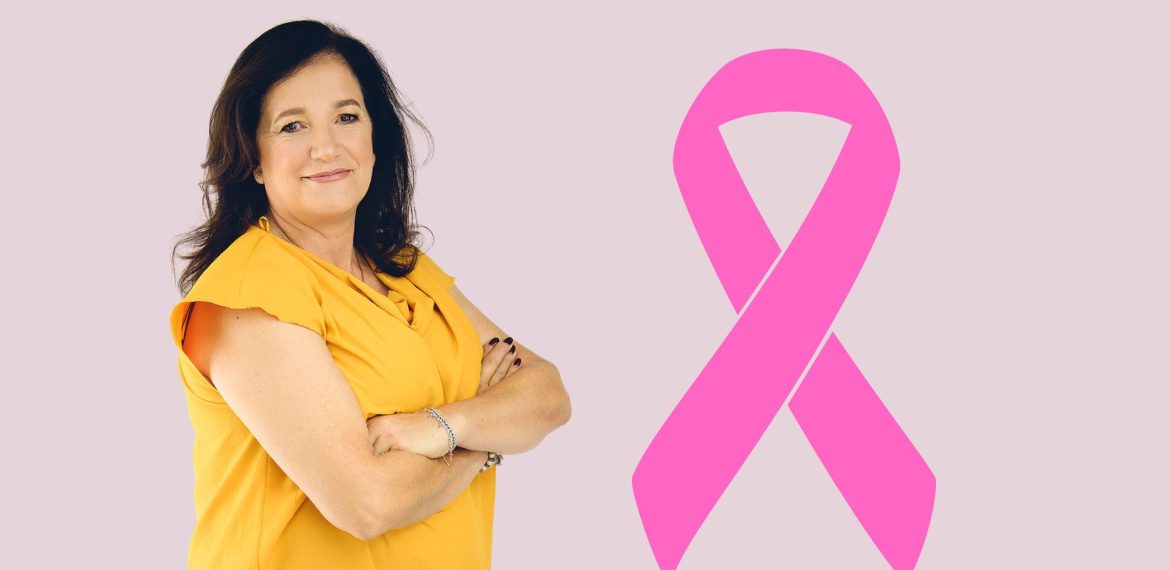 A woman in a yellow top stands with arms crossed, smiling, next to a large pink Shona breast cancer awareness ribbon on a light purple background.