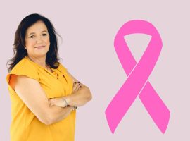 A woman in a yellow top stands with arms crossed, smiling, next to a large pink Shona breast cancer awareness ribbon on a light purple background.