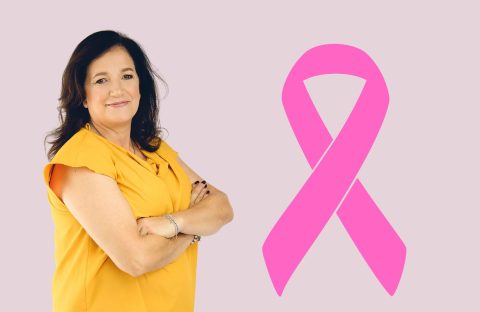 A woman in a yellow top stands with arms crossed, smiling, next to a large pink Shona breast cancer awareness ribbon on a light purple background.