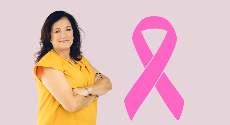 A woman in a yellow top stands with arms crossed, smiling, next to a large pink Shona breast cancer awareness ribbon on a light purple background.