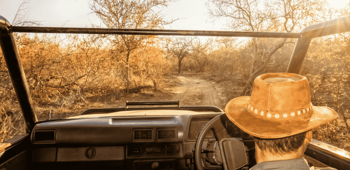 A person wearing a brown hat drives an open-top vehicle along a dirt road through a dry, sunlit landscape with sparse trees and golden foliage, a winter blanket tossed across the back seat for warmth on cooler evenings.