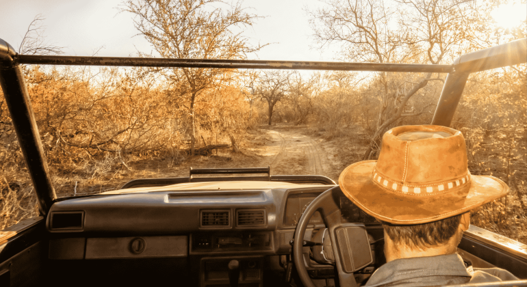 A person wearing a brown hat drives an open-top vehicle along a dirt road through a dry, sunlit landscape with sparse trees and golden foliage, a winter blanket tossed across the back seat for warmth on cooler evenings.