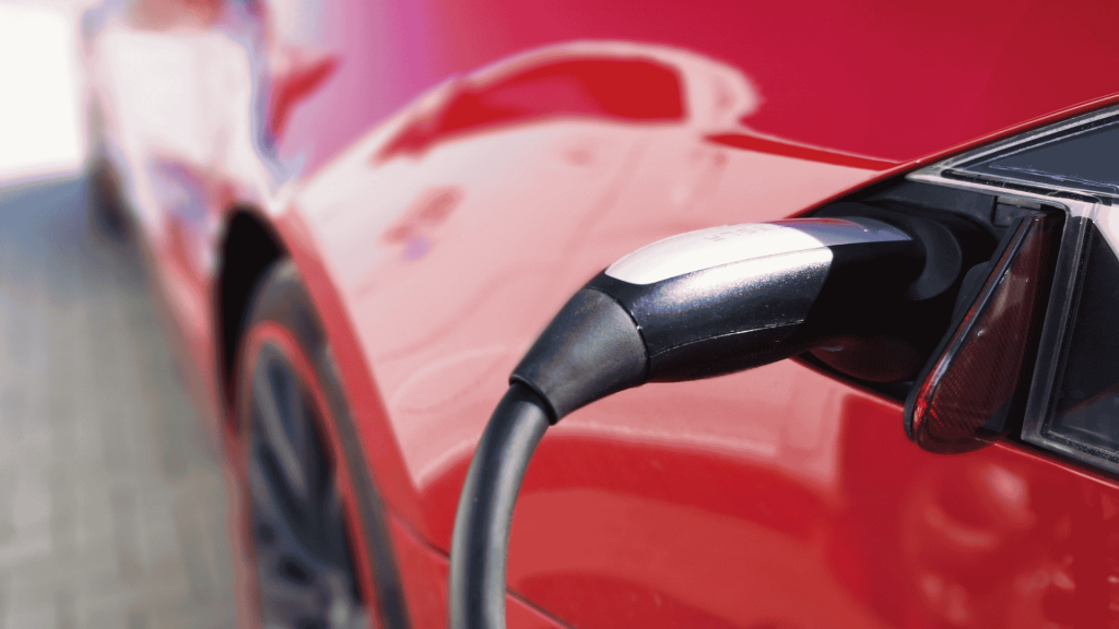 Close-up of an electric vehicle charging, showing the charging cable plugged into the port of a shiny red car, as a light winter blanket of snow dusts the surroundings.