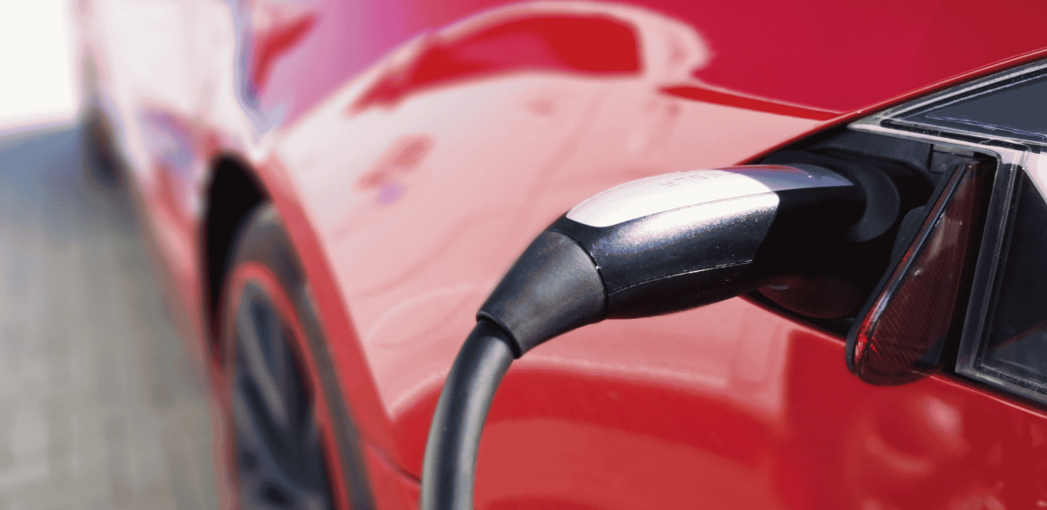 Close-up of an electric vehicle charging, showing the charging cable plugged into the port of a shiny red car, as a light winter blanket of snow dusts the surroundings.