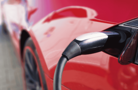 Close-up of an electric vehicle charging, showing the charging cable plugged into the port of a shiny red car, as a light winter blanket of snow dusts the surroundings.