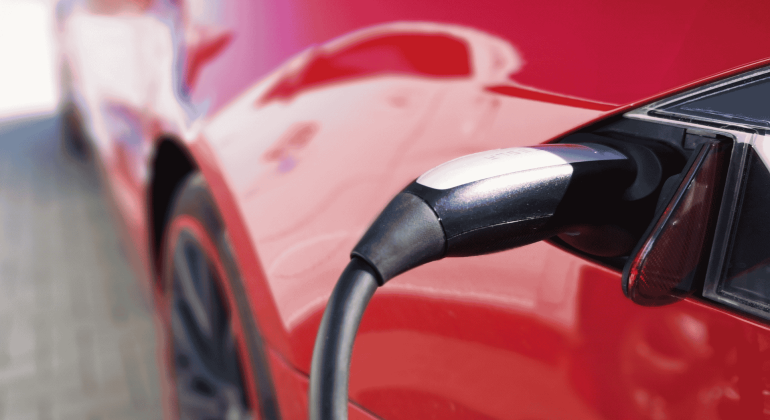 Close-up of an electric vehicle charging, showing the charging cable plugged into the port of a shiny red car, as a light winter blanket of snow dusts the surroundings.