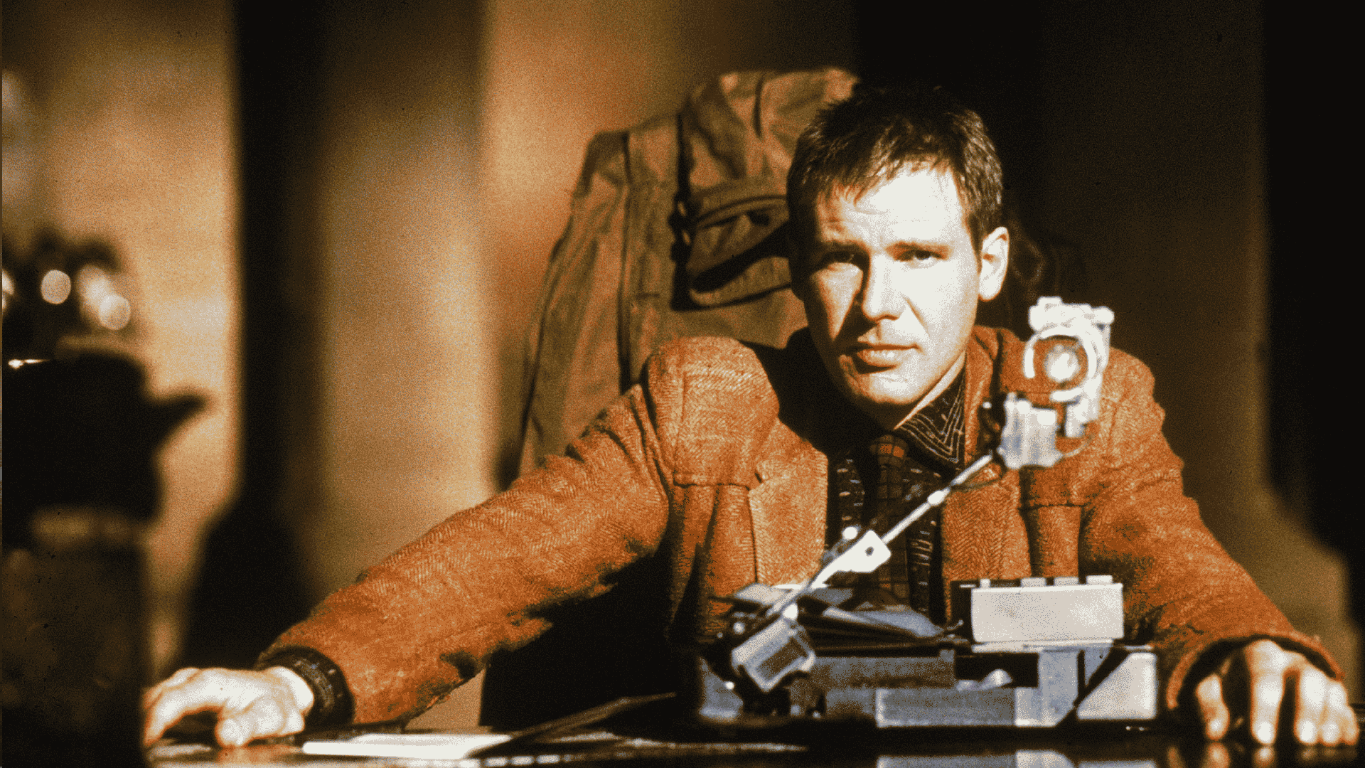 A man in a brown jacket sits at a desk with a futuristic machine in front of him, illuminated by warm light.