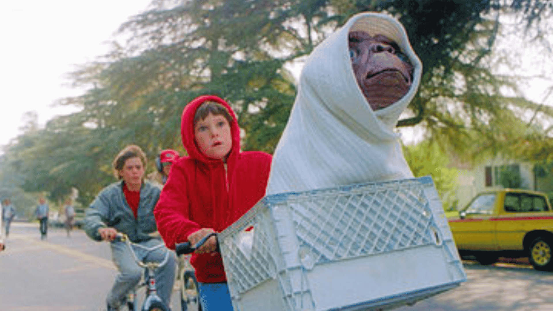 A boy in a red hoodie rides a bicycle with an alien wrapped in a white blanket in the front basket; another child follows on a bike.