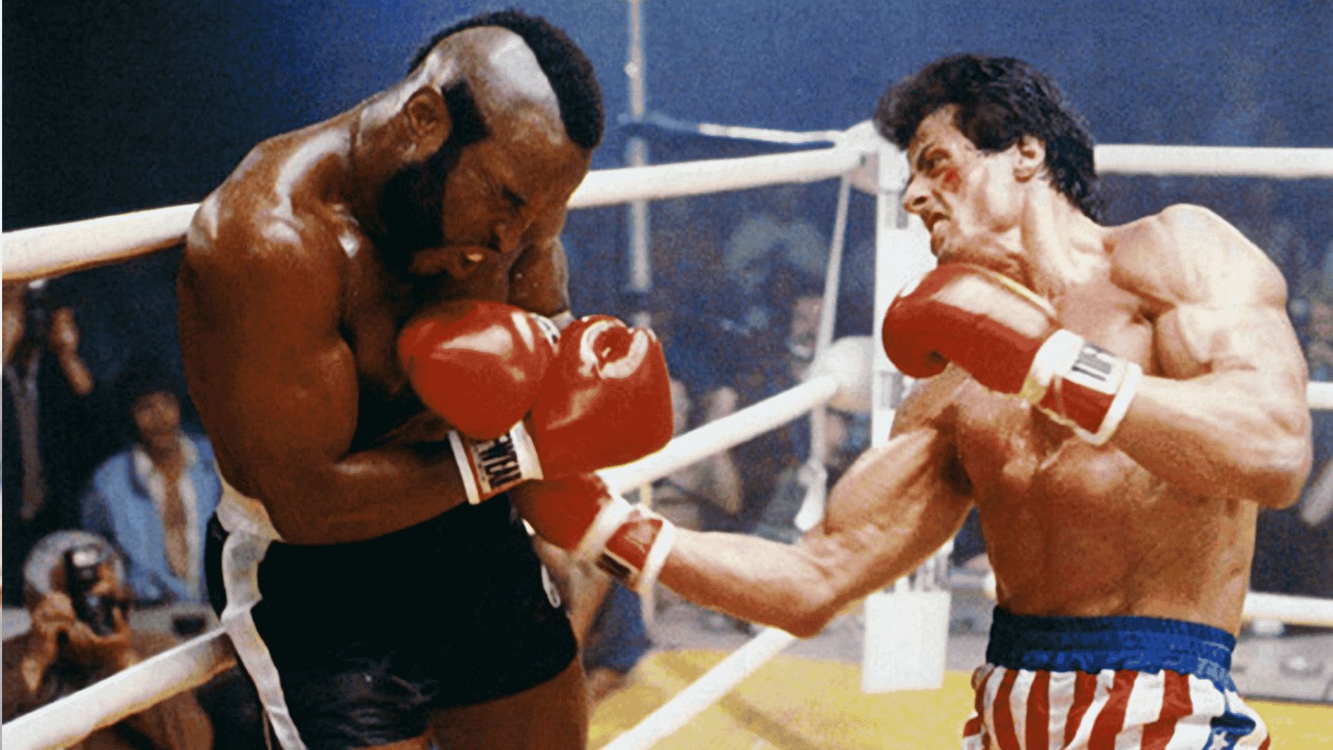 A boxer in red, white, and blue shorts punches his opponent, who is wearing red gloves and dodging defensively in a boxing ring. Spectators watch the intense match.