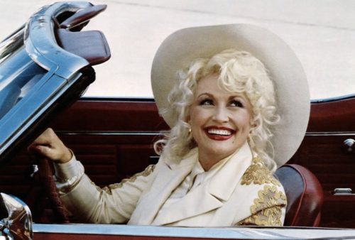 Smiling woman with blonde curly hair and a wide-brimmed white hat sits in a red convertible, wearing a white jacket with gold embroidery, looking up and to the side.