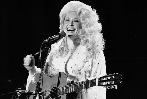 A woman with big blonde hair smiles while playing an acoustic guitar and singing into a microphone on stage, wearing a jeweled outfit.