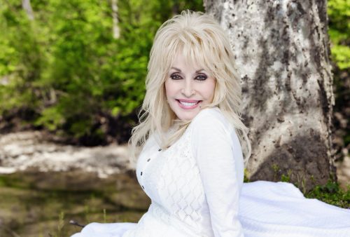 A woman with long blonde hair in a white outfit sits on a blanket outdoors, smiling with a forest and tree trunk in the background.