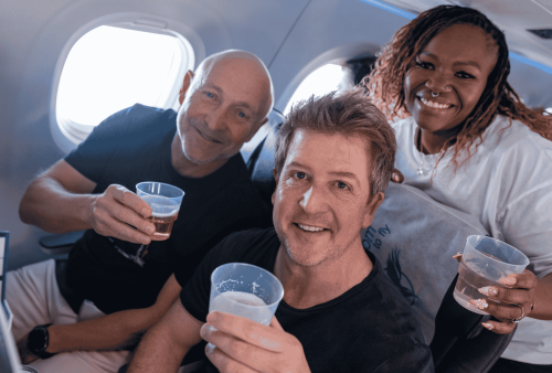 Three adults sitting on an airplane, smiling and holding plastic cups with drinks, appear to be celebrating or enjoying a cheerful moment together during their flight, while a cozy winter blanket is draped across their laps, adding warmth and comfort to the scene.