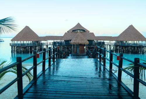 A wooden walkway leads to a thatched-roof structure on stilts over turquoise water, with palm leaves framing the scene and a sandy beach in the foreground&mdash;a perfect escape where even in winter you could trade your blanket for the gentle warmth of tropical sun.