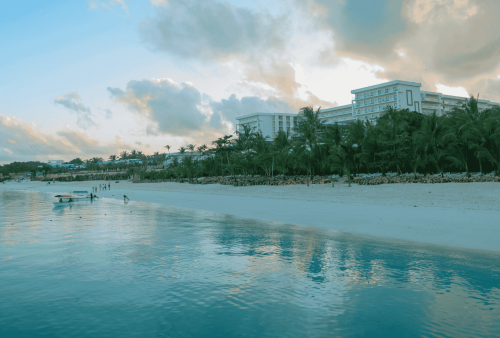 A calm beach scene with clear water, white sand, palm trees, and several large resort buildings in the background under a cloudy sky. A few people and a small boat are near the shore, while the sky above appears to wrap the beach in a soft winter blanket of clouds.