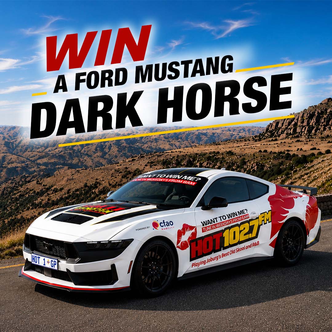 A white Ford Mustang with HOT 1027 FM branding is parked on a scenic mountain road. Large text above reads, WIN A MUSTANG DARK HORSE. Experience the thrill of the Mustang Dark Horse with every mile.