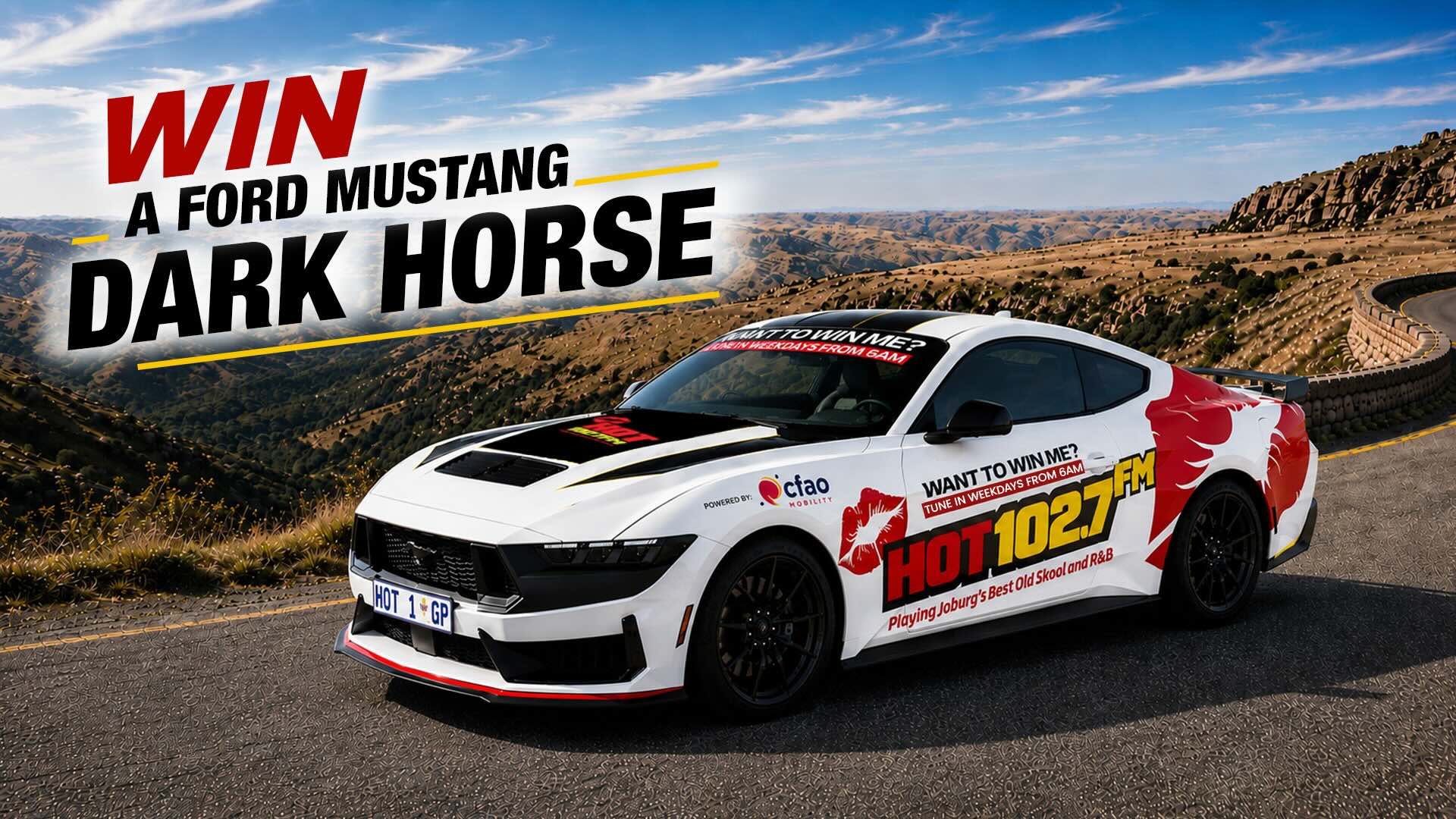 A white Ford Mustang Dark Horse with HOT 1027 FM branding is parked on a mountain road. Text reads: WIN A FORD MUSTANG DARK HORSE. Scenic mountains and blue sky are in the background.
