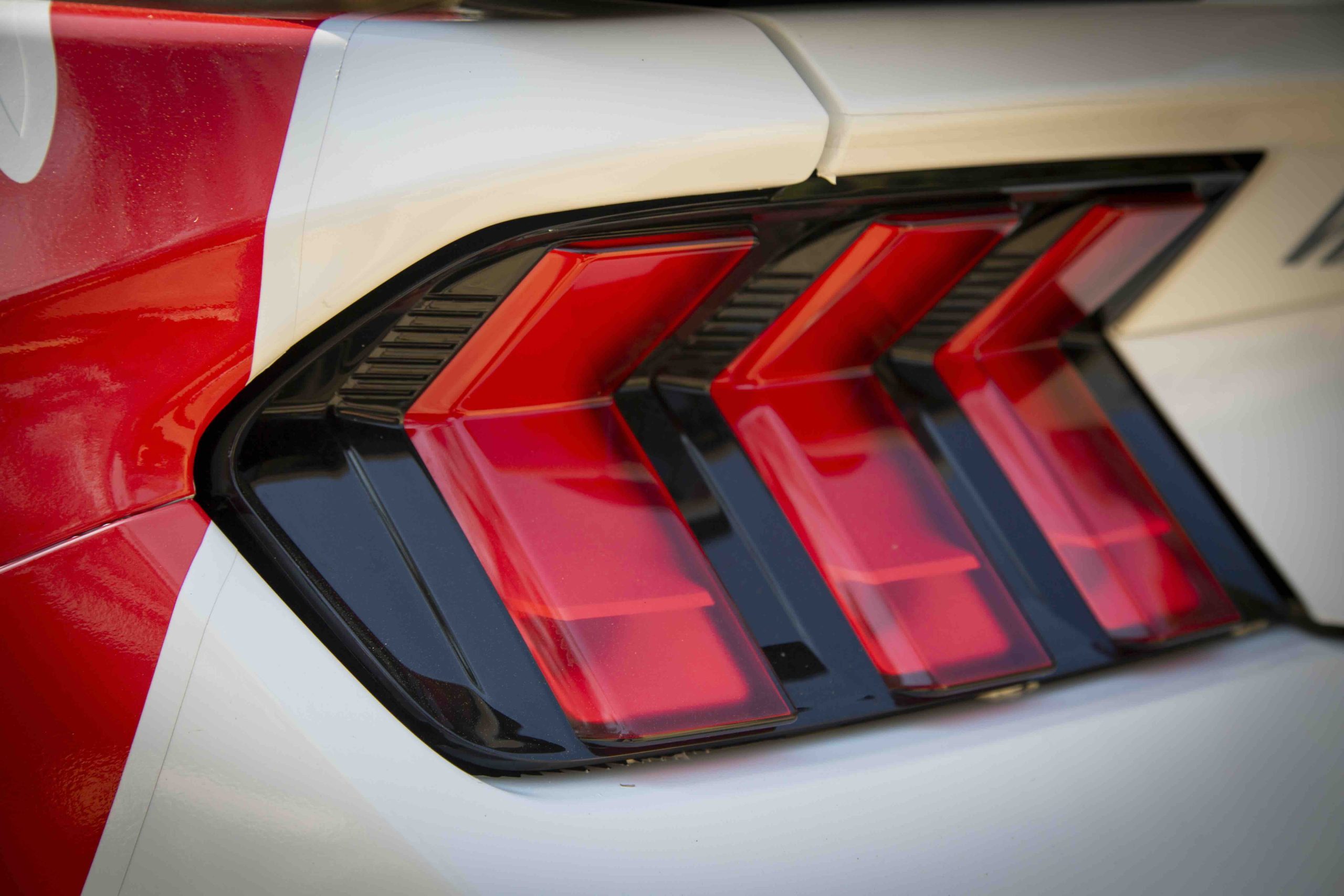 Close-up of the rear taillight of a red and white Mustang Dark Horse, showcasing three red, angular light sections with black trim.