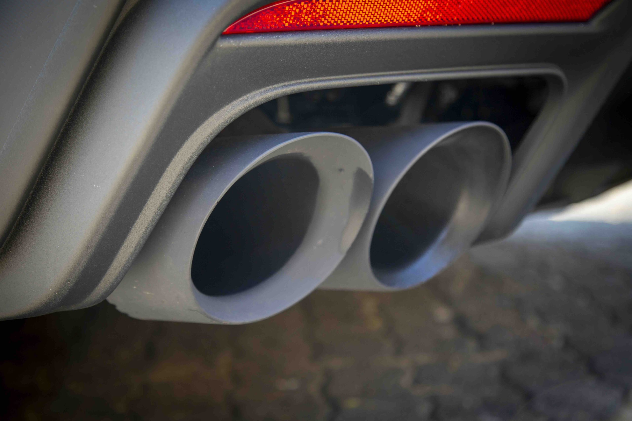 Close-up of a Mustang Dark Horse&rsquo;s dual exhaust pipes with a red reflector above them, mounted on a dark-colored bumper. The surface below is paved with stone.