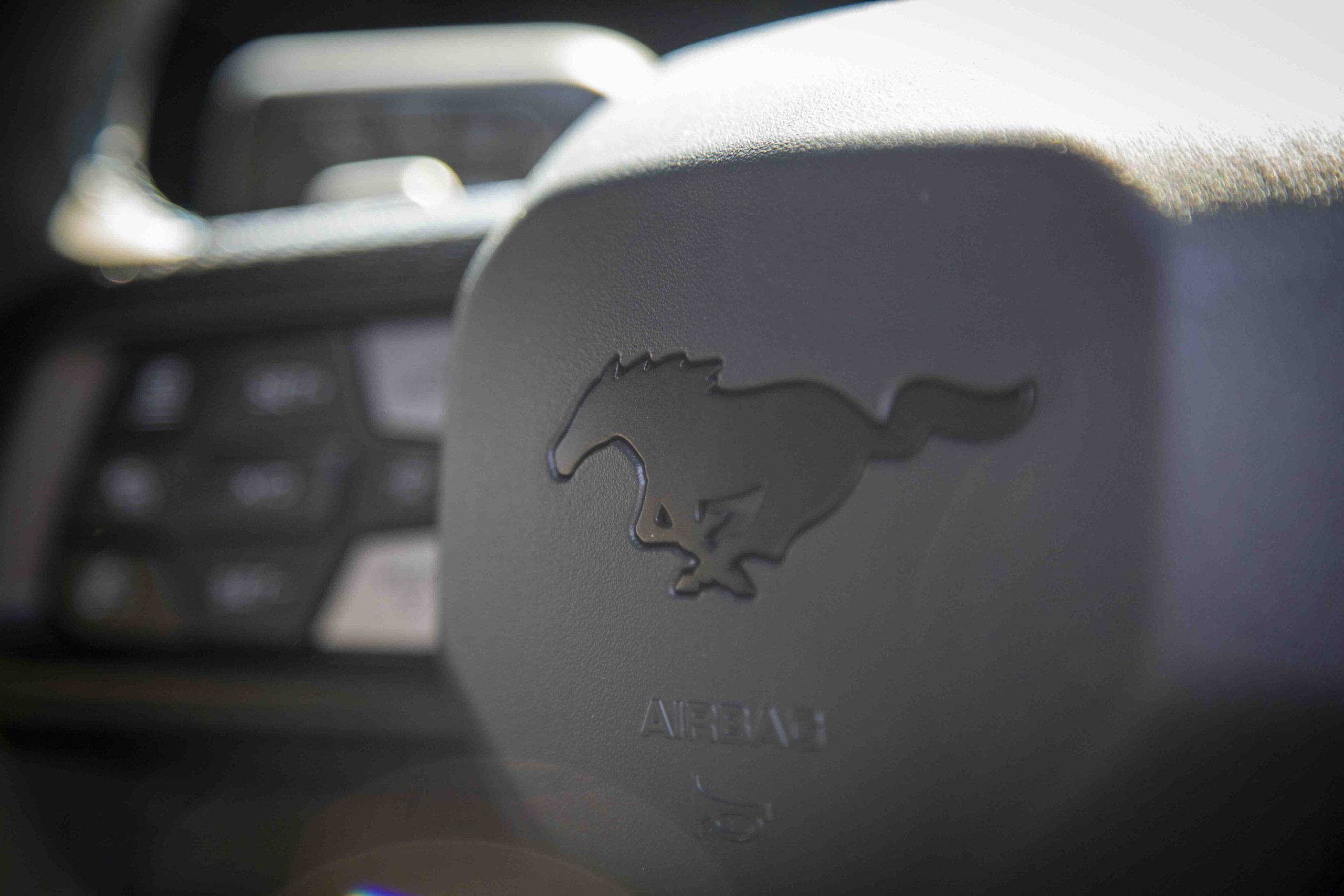 Close-up of a black steering wheel featuring an embossed galloping horse logo, with the word AIRBAG below it&mdash;likely from a Mustang Dark Horse. Blurred dashboard controls are visible in the background.