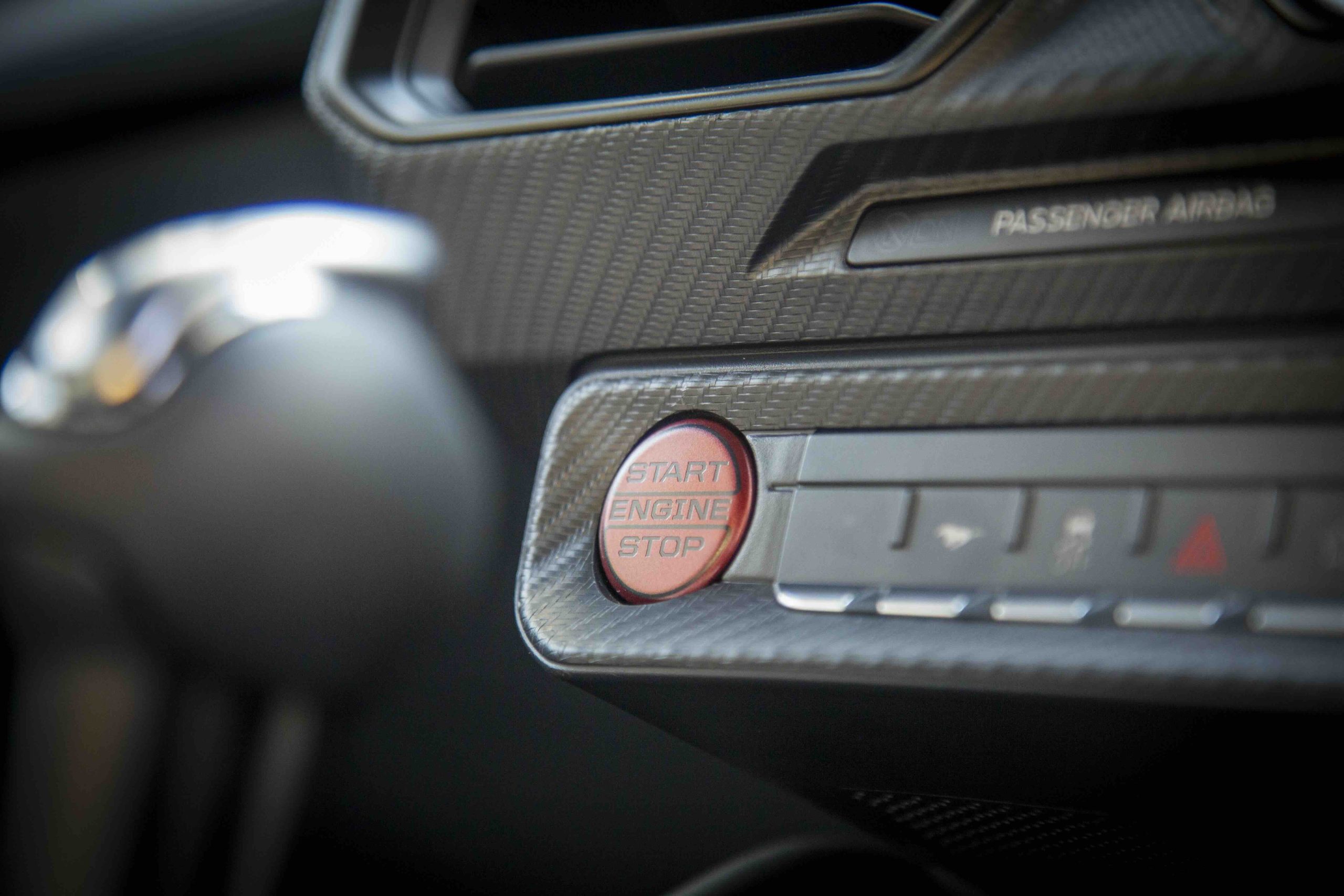 Close-up of a red Start Engine Stop button on a Mustang Dark Horse car dashboard, with nearby controls and Passenger Airbag label visible.