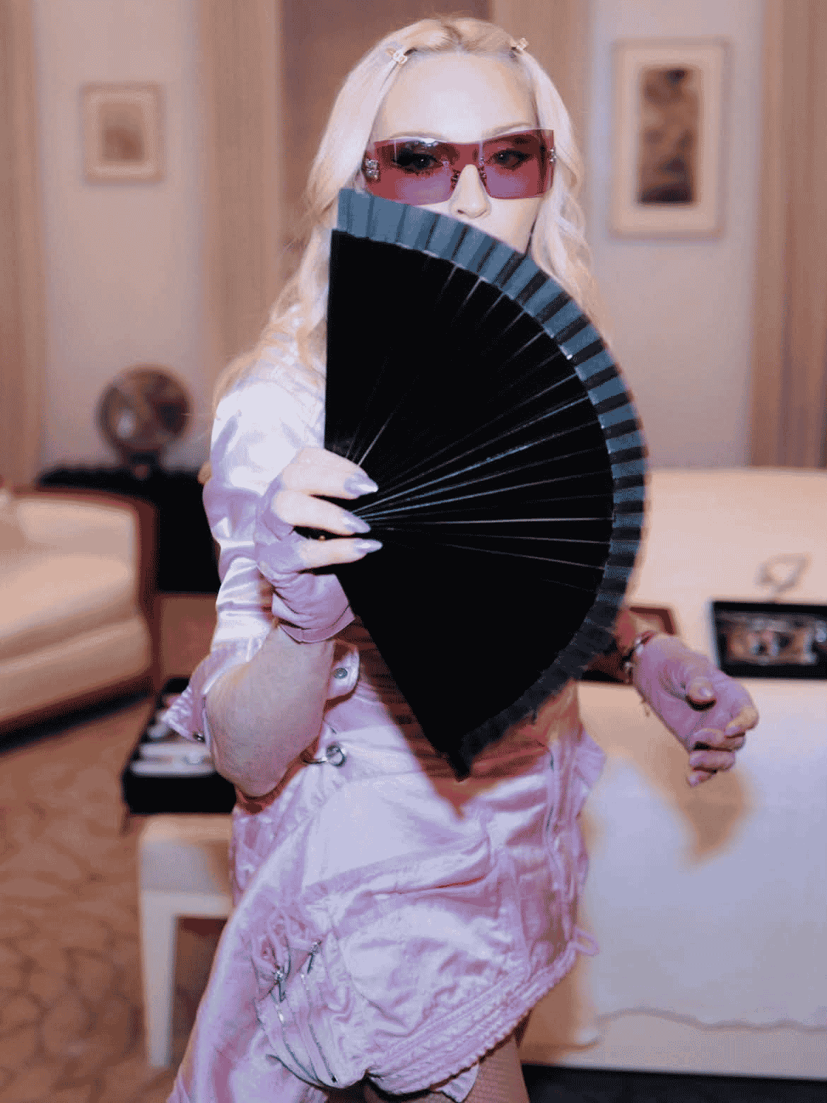 Person in pink outfit and sunglasses holds a black fan partially covering their face, standing in a softly lit room with a couch, table, and framed wall art in the background.