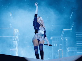 A performer stands on stage with one arm raised, holding a microphone, in front of a blue-lit background with cityscape elements and fog effects.