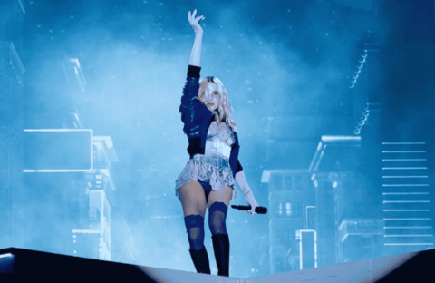A performer stands on stage with one arm raised, holding a microphone, in front of a blue-lit background with cityscape elements and fog effects.