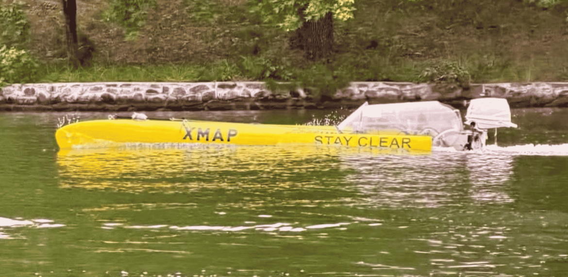 A long yellow boat labeled XMAP STAY CLEAR travels on a green river near a stone-lined shore, with grass and trees in the background, evoking a scene as vibrant and spirited as a Dolly Parton country song.