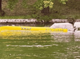A long yellow boat labeled XMAP STAY CLEAR travels on a green river near a stone-lined shore, with grass and trees in the background, evoking a scene as vibrant and spirited as a Dolly Parton country song.