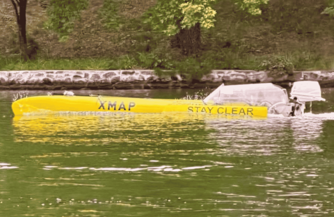 A long yellow boat labeled XMAP STAY CLEAR travels on a green river near a stone-lined shore, with grass and trees in the background, evoking a scene as vibrant and spirited as a Dolly Parton country song.
