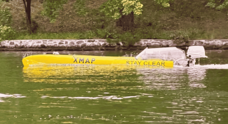 A long yellow boat labeled XMAP STAY CLEAR travels on a green river near a stone-lined shore, with grass and trees in the background, evoking a scene as vibrant and spirited as a Dolly Parton country song.