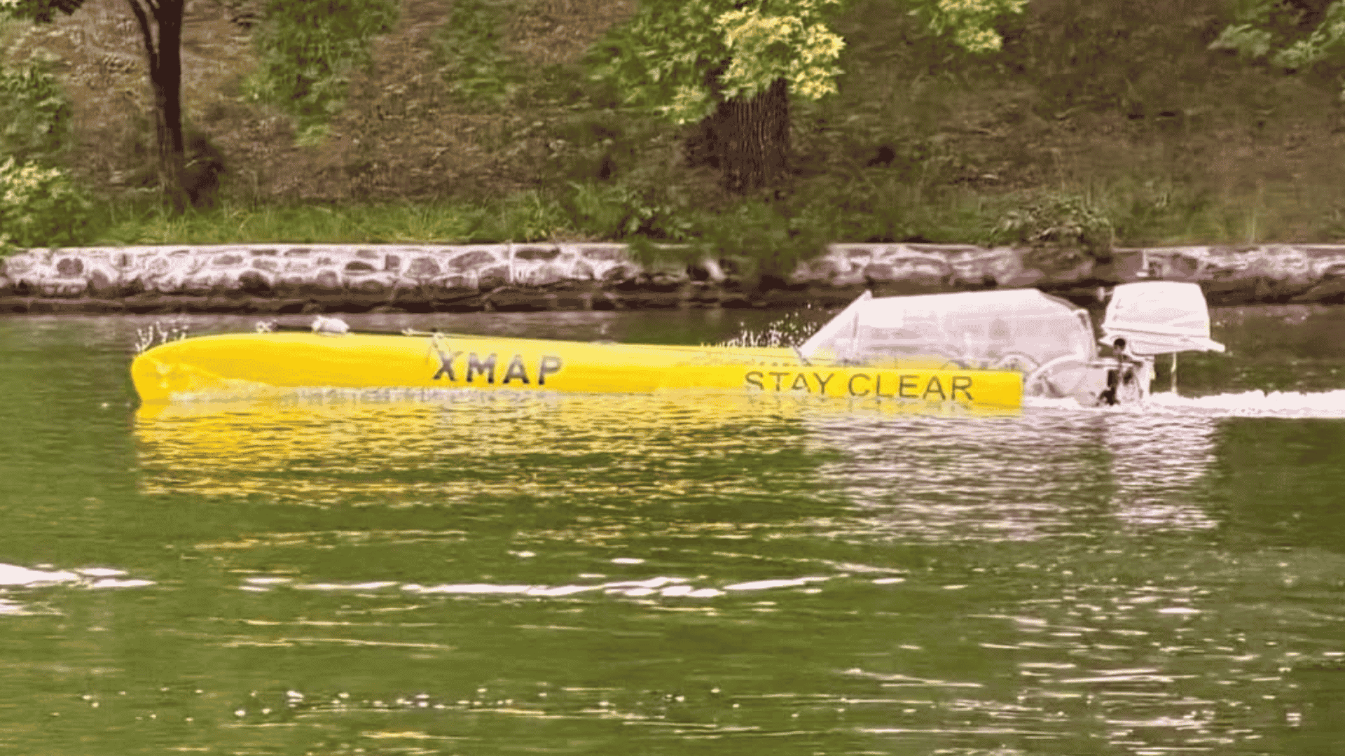 A long yellow boat labeled XMAP STAY CLEAR travels on a green river near a stone-lined shore, with grass and trees in the background, evoking a scene as vibrant and spirited as a Dolly Parton country song.