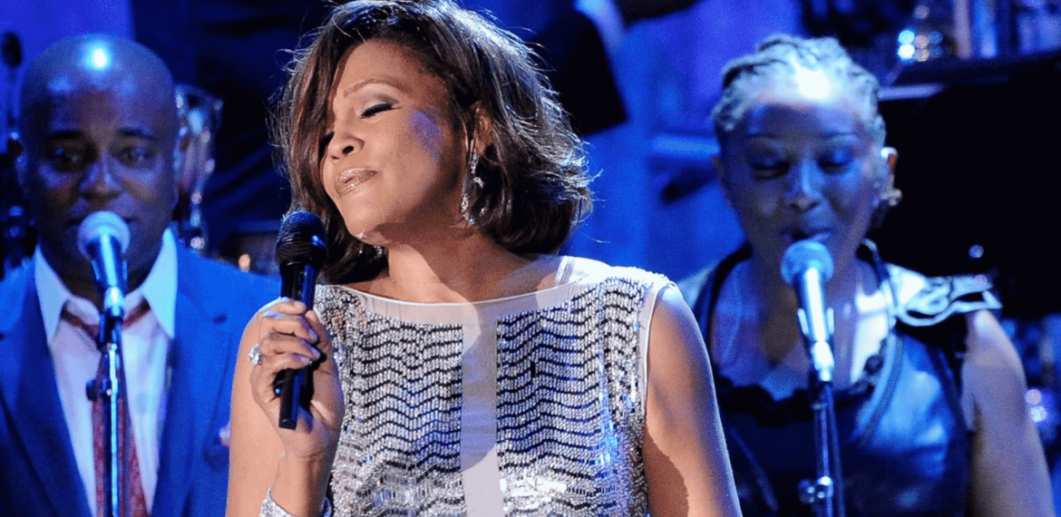 A woman in a sparkly dress sings into a microphone on stage, eyes closed, with two backup singers and a blue-lit background.