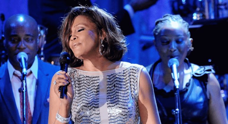 A woman in a sparkly dress sings into a microphone on stage, eyes closed, with two backup singers and a blue-lit background.