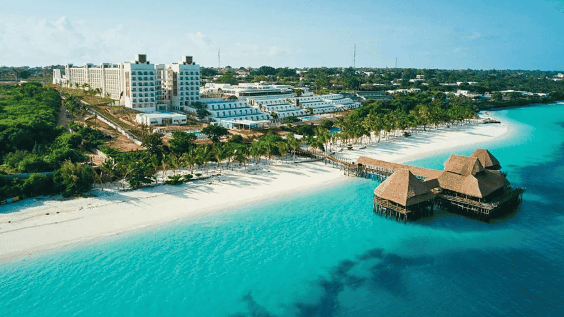 Aerial view of a resort with modern buildings, palm trees, white sandy beach, and turquoise ocean&mdash;an exclusive oasis perfect for Amex members. A wooden pier with thatched-roof huts extends over the water, offering a luxurious escape.