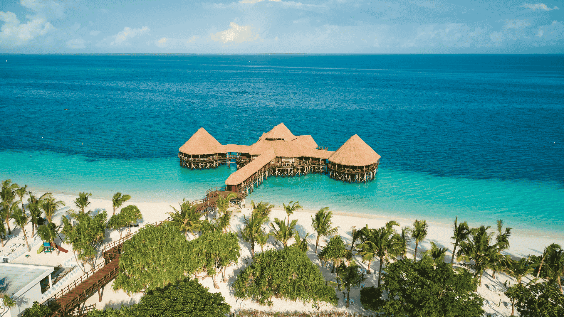 Aerial view of a wooden pier with thatched-roof huts, reminiscent of an Amex travel destination, extending over turquoise ocean water from a white sandy beach lined with palm trees.