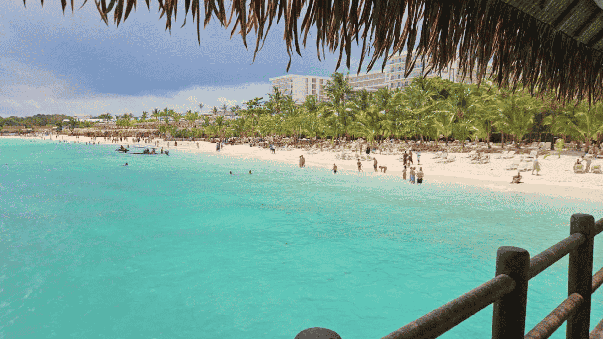 View of a tropical beach with turquoise water, people swimming, palm trees, and a white hotel in the background, seen from under a thatched roof—a perfect escape from the winter blanket that covers much of the world.