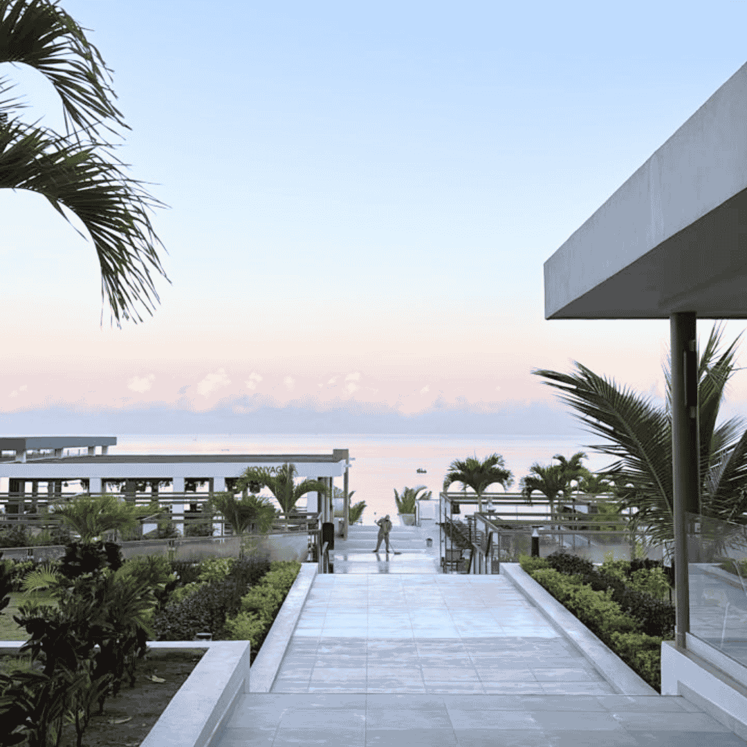 A modern resort walkway with palm trees leads to the ocean at sunrise or sunset. Wrapped in a cozy winter blanket, a person walks toward the beach, surrounded by greenery and contemporary architecture under a clear sky.