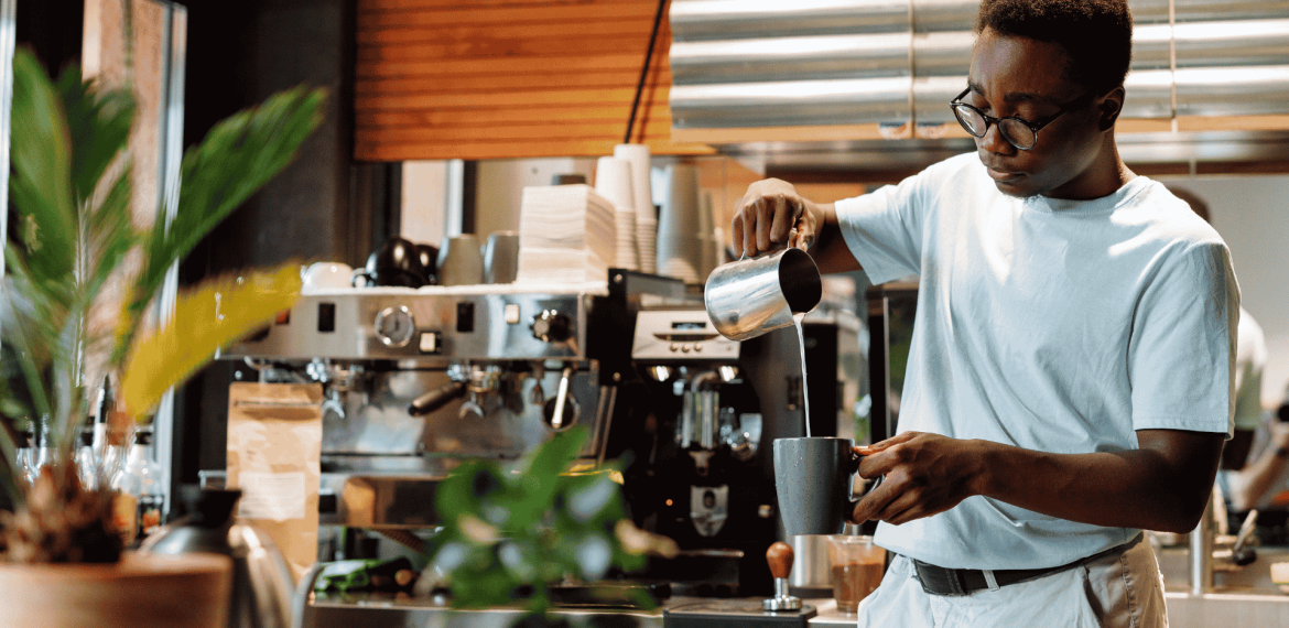 A barista wearing glasses pours steamed milk into a cup at a coffee shop, standing in front of an espresso machine and surrounded by plants and coffee equipment, while wrapped in the cozy comfort of a winter blanket.