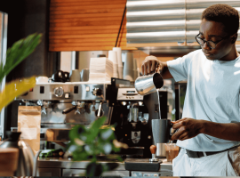 A barista wearing glasses pours steamed milk into a cup at a coffee shop, standing in front of an espresso machine and surrounded by plants and coffee equipment, while wrapped in the cozy comfort of a winter blanket.